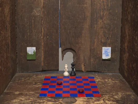 A detail photograph depicting the inside of a wooden bathtub titled upright. A red and blue painted checkerboard pattern is painted onto the wooden base where two red stones are placed on blue squares within the checkerboard and a white knight chess piece and a black queen chess piece are placed in the back overlooking the checkerboard. In the background, two miniature landscape paintings hang.