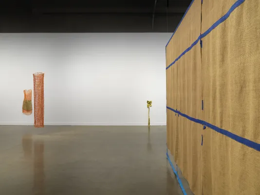 On the left, two cylindrical orange net-like sculptures are displayed. One is much smaller than the other; the smaller one incorporates an inner green component. On the far right, a large brown tapestry hangs from the ceiling. Blue fiber strips grid the piece with some of the strips dangling onto the floor. In the background on the right, a small, yellow caution tape fiber sculpture is bunched up dangling off the wall and onto the floor.