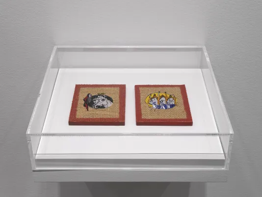 A plexiglass case hangs off of a white wall. Inside the case, two burlap-like square artworks are adorned with embroidered details. The left shows the decorated head of a horse, while the left is more ambiguous but has a similar medieval flavor. Each square has a border of red wax around the piece.
