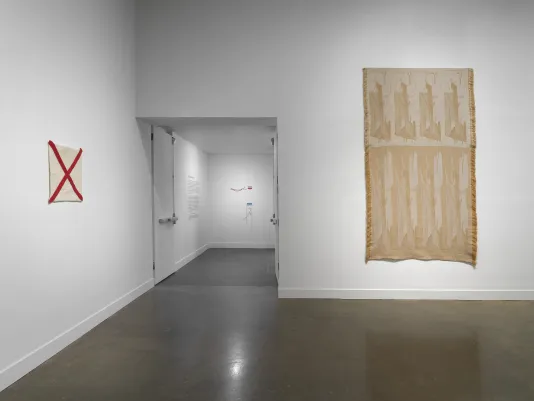 The entryway to a gallery is shown from the viewpoint of someone inside. To its left, an off-white fiber square with a red X across the piece hangs on the wall. To its right, a large, brown and beige tapestry with fringe edges hangs on the wall. In the back of the entrance way, a small two-part fiber artwork hangs on the wall.