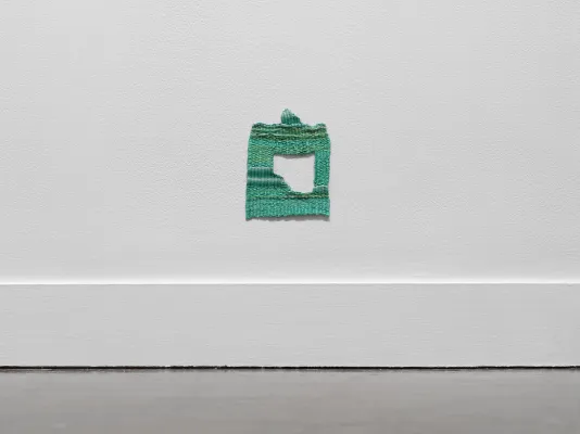 A small green square shaped weaving hangs on the wall close to the floor. A hole in its center, shaped approximately like a square with a missing corner, reveals the white wall behind the piece.