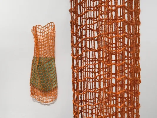 A close-up view of a woven netlike structure of a hanging textile artwork. The artwork forms a kind of dense cylinder, with several overlapping layers of material. On the left of the image, another orange netlike artwork hangs on the wall encasing a smaller green fiber cylinder inside of it.