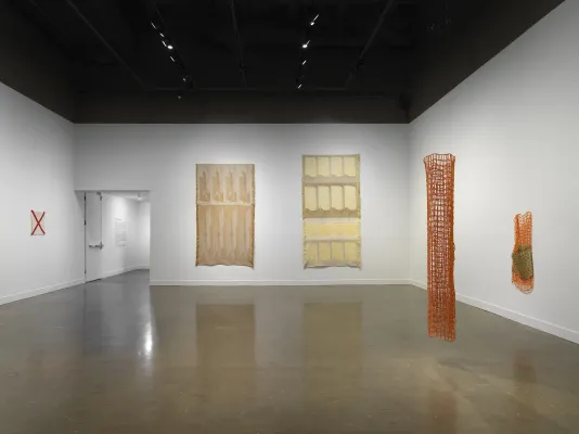 A white gallery space with a reflective cement floor. On the right, two netlike orange sculptures are on display with one hanging off the wall and one suspended from the ceiling. In the back, two large, brown and beige tapestries with fringe edges and a repetitive pattern hang on the wall. On the left, an off-white fiber square with a red X across the piece hangs on the wall next to the entrance to the gallery.