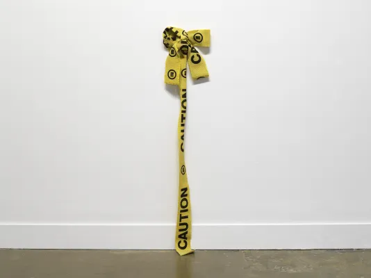 A yellow woven, caution tape fiber artwork is bunched up in a bow-like form dangling off the white wall onto the cement floor.