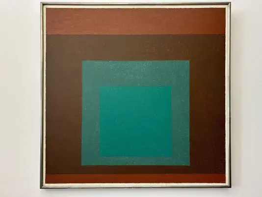 Square painting with a silver frame on a white wall of a brown background with a smaller turquoise square in the center and a lighter brown stripe across the top.