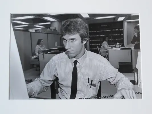 Black and white photograph of a man intensely focused working in an office, wearing a long dark colored tie and two pencils in his shirt pocket.
