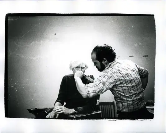 Black and white photograph of two men, one is seated with a dark colored long sleeve shirt and white hair and the other is standing with his hand raised in front of the other mans face.