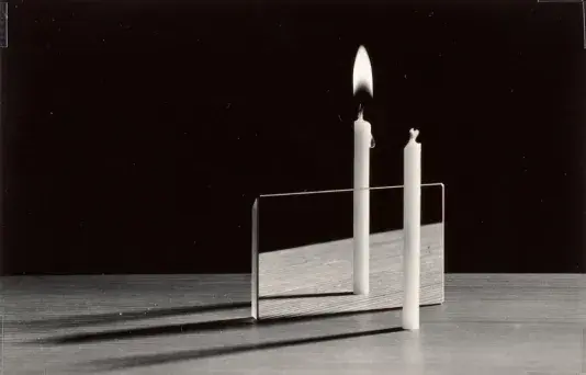 Black and white photograph of two candles on a table one is lit and the other is not. The candle with a flame appears behind the mirror and the unlit candle is positioned in front.