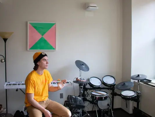 A man wearing a yellow shit sitting in front of an artwork with a black frame on the white wall behind. There is a drum set on the right side.