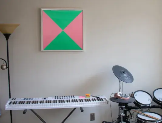Artwork with a black frame hangs on a wall above a electric keyboard. There is a drum set on the right side and a tall lamp to the left.