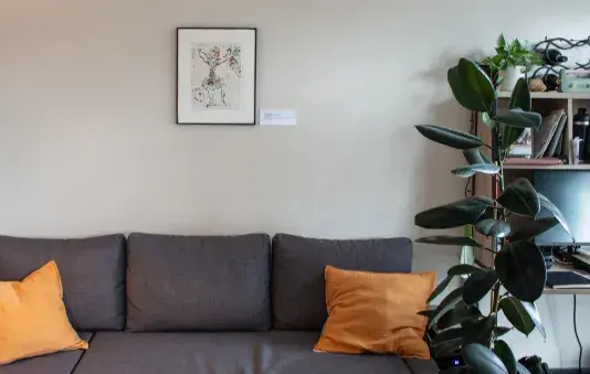 A work of art with a black frame mounted on the white wall above a gray couch with two yellow pillows. Part of a large plant is in view on the right side.