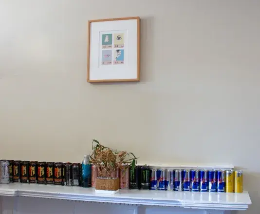 A white mantle with a row of energy drink cans in a line on top. A framed artwork hangs on the wall above.
