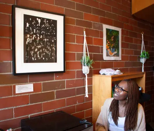 A woman with glasses wearing a beige cardigan to the right of an artwork with a black frame hung on a brick wall.