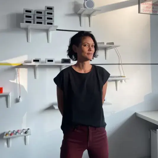 Woman with short dark hair, wearing a black t-shirt and burgundy pants, stands with her arms behind her back in front of a bright white wall with shelves mounted in various heights.