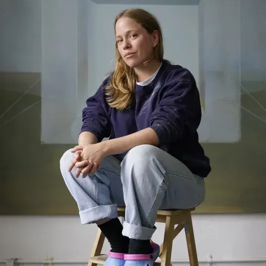 Woman wearing light jeans and a navy crewneck with vibrant periwinkle crocs sits on a wooden stool in the middle of an open art studio.