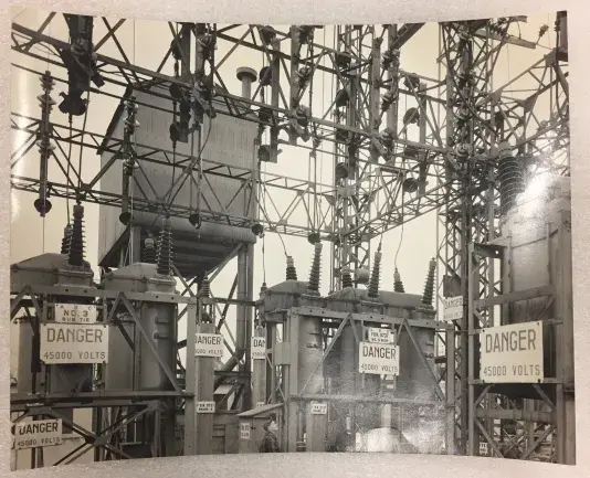 Black and white photograph of a collection of transformers with nine danger signs hung on various heights.