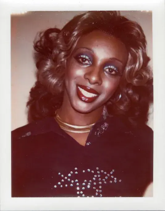Photograph of a woman with bright white smile and voluminous curly hair, wearing blue eyeshadow, gold necklaces and gold hoop earrings..