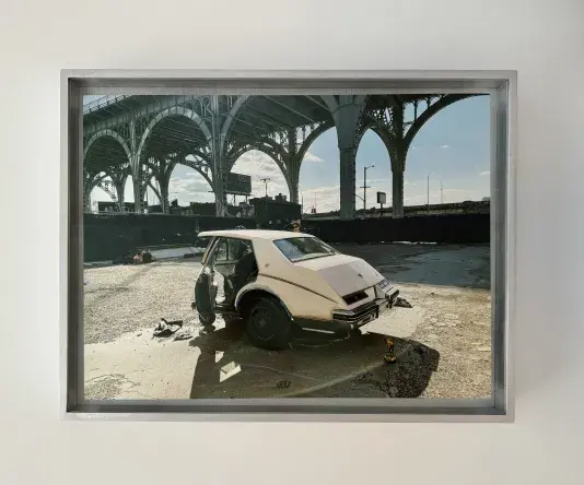 Colored photograph in a silver frame of the back half of a white car parked in an open dirt lot with a tall metal bridge in the background.