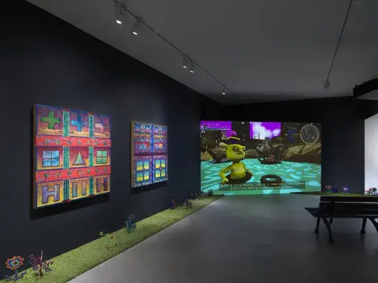 A dark, black-walled gallery space with artworks on display. On the left wall, two‬ ‭ paintings hang in a row depicting bright, geometric forms. In the back of the gallery, a‬ ‭ large screen illuminates the space displaying digitally rendered footage of a green‬ ‭ creature in water. A park bench faces the screen. A strip of astroturf lines the floor‬ ‭ around the gallery, with painted wooden flowers emerging from it.