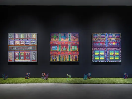 Three square paintings hang in a row on a black wall. The paintings depict‬ ‭ bright, geometric forms. A row of astroturf lines the floor against the wall, with small‬ ‭painted wooden plants emerging from it.‬ ‭