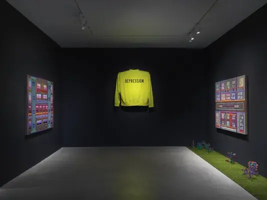 An art gallery with dim lighting showcases three artworks on the walls. In the‬ ‭ center, a bright neon yellow sweater with the word ‘Depression’ written across it is‬ ‭ displayed, elevated off the wall. On the left and right walls, two paintings with bright‬ ‭ geometric forms hang across from one another. A strip of astroturf on the floor lines the‬ ‭ right wall with small painted wooden plants emerging from it.‬