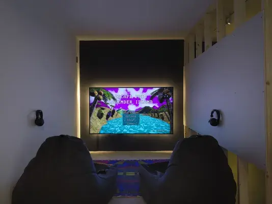 A backlit, glowing TV screen hangs on the wall close to the floor. A start menu‬ ‭ depicting a digitally rendered island scene for the videogame‬‭ Escape from Lavender‬ ‭ Island‬‭ is displayed on the TV. A pair of headphones hang off both the left and right‬ ‭ walls. Two soft chairs are placed on the floor over a colorful rug in front of the screen.‬