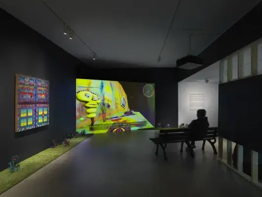 A dark, black-walled gallery space with artworks on display. On the left wall‬ ‭ hangs a painting depicting bright, geometric forms. In the back of the gallery, a large‬ ‭ screen illuminates the space displaying digitally rendered footage of a bright colored,‬ ‭ cartoon-like room. A woman sits on a park bench facing the screen. A strip of astroturf‬ ‭ lines the floor around the gallery, with painted wooden flowers emerging from it.‬