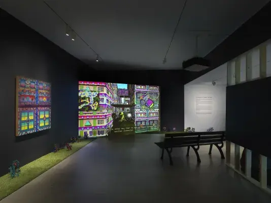 A dark, black-walled gallery space with artworks on display. On the left wall‬ ‭ hangs a painting depicting bright, geometric forms. The same forms in the painting are‬ ‭ echoed in the digitally rendered city scene that is projected on a large screen in the‬ ‭ corner of the room. A park bench in the center of the room faces the screen. A strip of‬ ‭ astroturf lines the floor around the gallery, with painted wooden flowers emerging from‬ ‭it.‬