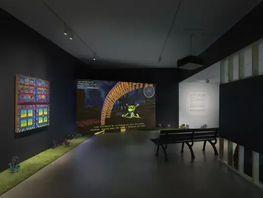 A dark, black-walled gallery space with artworks on display. On the left wall‬ ‭ hangs a painting depicting bright, geometric forms. In the back of the gallery, a large‬ ‭ screen illuminates the space displaying digitally rendered footage of a green creature‬ ‭ reading a book under an orange archway overlooking a city. A park bench faces the‬ ‭ screen. A strip of astroturf lines the floor around the gallery, with painted wooden‬ ‭flowers emerging from it.‬ ‭