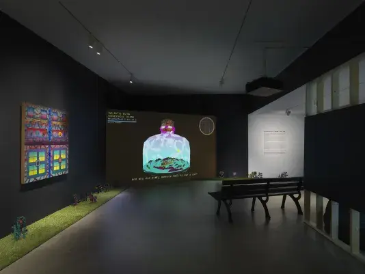A dark, black-walled gallery space with artworks on display. On the left wall‬ ‭ hangs a painting depicting bright, geometric forms. In the back of the gallery, a large‬ ‭ screen illuminates the space displaying digitally rendered footage of a large bottle with‬ ‭ an island inside of it. A park bench faces the screen. A strip of astroturf lines the floor‬ ‭ around the gallery, with painted wooden flowers emerging from it.‬