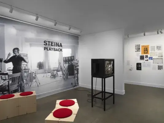 A black-and-white photograph printed mural-size on a wall shows a woman‬ ‭ wearing headphones. She is located in a studio full of technical equipment: tripods,‬ ‭video monitors, wiring. The photograph is overlaid with text indicating the exhibition title:‬ ‭ “Steina: Playback.” The gallery space also includes a boxy video monitor and wooden‬ ‭ seating. In the background on the right, posters and other documents are pasted on the‬ ‭wall.‬ ‭