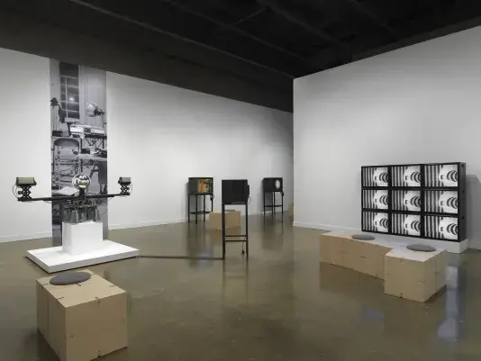 A gallery with several video works. At left is a sculptural apparatus with two‬ ‭ cameras on the ends of a single rotating arm, each pointed inwards at a central‬ ‭ mirrored sphere. At right is a grid of nine monitors, all showing the same abstract‬ ‭ black-and-white image. In the middle, three boxy video monitors on metal stands are‬ ‭visible in the background.‬ ‭