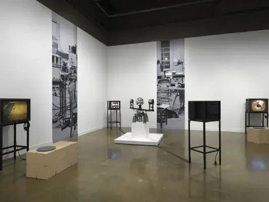 In the center, a sculptural apparatus comprises two cameras on the ends of a‬ ‭ single rotating arm, each pointed inwards at a central mirrored sphere. Feeds from the‬ ‭ cameras are routed to two monitors in the space. On the walls nearly, strips of a‬ ‭ photograph of a studio are pasted on the wall at large scale. On either edge of the‬ ‭ image, video monitors display other works; the one on the right shows a body distorted‬ ‭around a circle, recalling the feed of the orb.‬ ‭