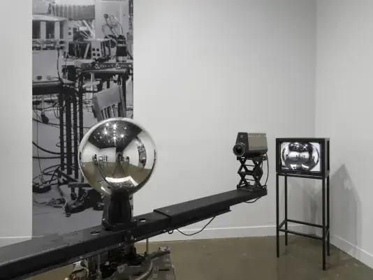 A mirrored sphere sits on top of a rotating central arm with cameras at each‬ ‭ end. In this close-up view, we see the details of the room within the orb: video monitors,‬ ‭ lights, the tripod taking the photo. In the corner behind the sculpture, a monitor shows a‬ ‭ feed of the orb, within which details of the room are also faintly visible. On the wall is a‬ ‭ narrow strip of a photograph of a studio space filled with technical equipment.