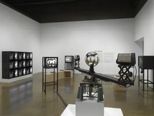 In the foreground, a sculptural apparatus comprises two cameras on the ends‬ ‭ of a single rotating arm, each pointed inwards at a central mirrored sphere. A feed from‬ ‭ the cameras are routed to a nearby monitor. In the background, other video monitors‬ ‭ are visible, including a grid of nine showing the same abstract black-and-white pattern,‬ ‭along with exhibition wall text.‬ ‭