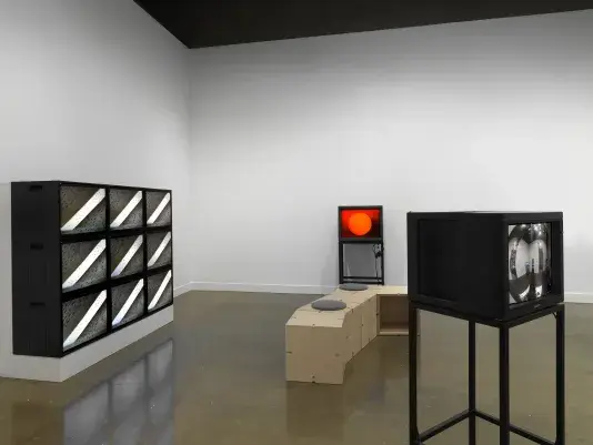 Boxy video monitors fill a gallery space. In the foreground at right, the monitor‬ ‭shows a gallery space as captured in the reflection of a mirrored orb. At left, a grid of‬ ‭nine showing the same abstract image: a white diagonal line against a dark gray‬ ‭ background. On the monitor in the background, a red circle is shown against a‬‭ background that fades from red to black.‬ ‭