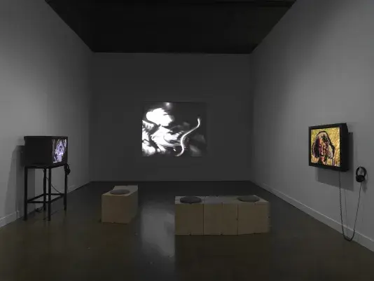 Three videos are shown in a gallery space. At center, a distorted image of a‬ ‭ person playing the violin is projected on the wall: the bow is curved and the person‬ ‭ indistinct, hinting at its electronic manipulation. At right, a flat monitor shows an image of‬ ‭ an older woman against a yellow background; portions of the background show through‬ ‭the face. At right, a boxy video monitor sits on a stand.‬ ‭