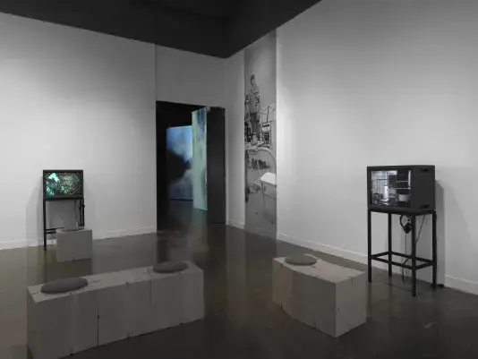 A gallery contains two boxy video monitors on metal stands, each with an‬ ‭ accompanying pair of headphones. The monitor on the left shows a color image that‬ ‭ could be the outline of trees against the sky. The right monitor shows a black-and-white‬ ‭ image of boxy shapes, possibly electronic equipment. A narrow slice of an image is‬ ‭ printed on the wall in the background, showing a woman in a studio space. Further‬ ‭ back, through a doorway, we glimpse a darker gallery that contains large-scale video‬ ‭