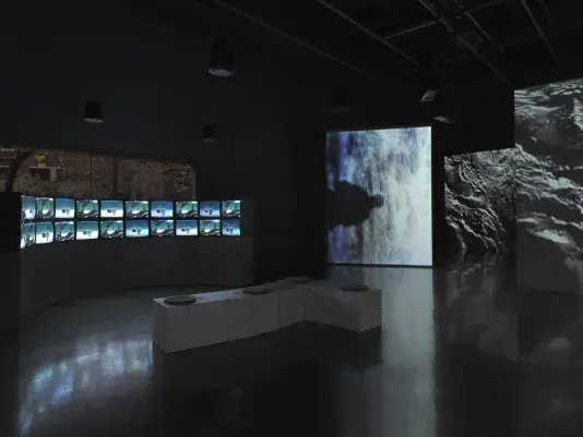 Two large-scale video installations occupy a darkened gallery. At left, a‬ ‭ double-stacked arc of boxy video monitors sits on a curved pedestal. In an alternating‬ ‭ pattern, the monitors show images of giant satellite dishes. At right, large‬ ‭ vertically-oriented screens display video projections of watery textures. One of the‬ ‭ screens shows a misty abstract surface with the shadow of a person’s head and torso‬ ‭jutting in from the right.‬ 