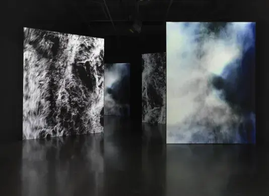 Four large vertically oriented video screens fill a dark gallery space. Two of‬ ‭ them (front left and back right) show a body of water seen from above, webbed with‬ ‭ whitecaps. The other two (front right and back left) show a misty abstract surface with‬ ‭ the shadow of a person’s head and torso jutting in from the left.