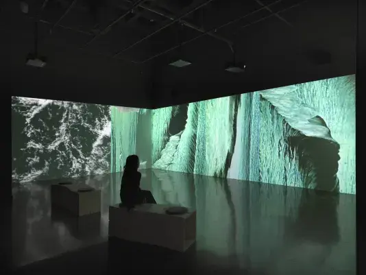 A continuous surface of projected video fills a wall and wraps around a corner‬ ‭ in a dark gallery. The images are liquid-like semi-abstractions of blue-green, white, and‬ ‭ black, recalling the surface of moving water. A person sits on one of two wooden‬ ‭ benches that are faintly visible in the foreground.‬ 