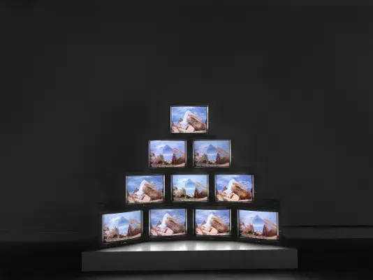 A pyramid of boxy monitors sits on a low pedestal in a dark space. The‬ ‭monitors display images of rocks against blue sky. Closer inspection reveals the images‬ ‭ have been electronically manipulated, overlaying multiple landscapes or sections of sky.‬