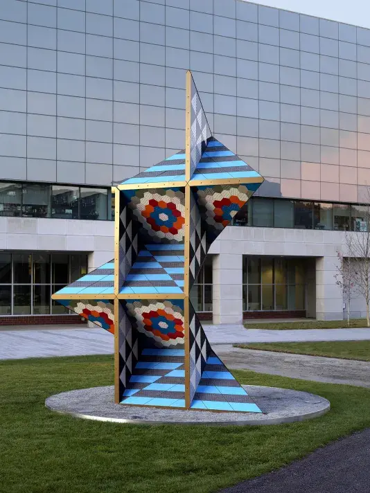 Large brightly colored geometric sculpture in the middle of a grass lawn in front of a concrete building with reflective blue glass windows.