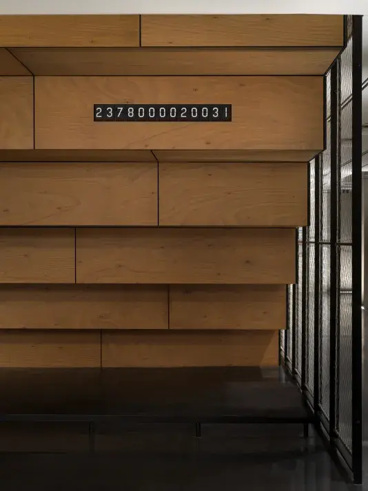 There is a black "ticker clock" with white numbers. The clock is placed on the underneath side of a wooden staircase.