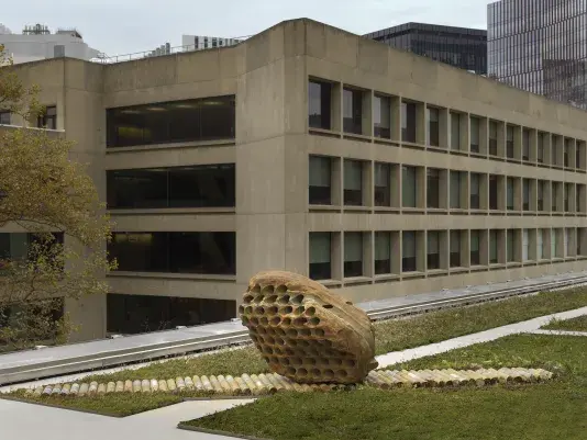 A large rock sculpture rests on a long strip of stone cylinders in a‬ ‭greenspace. The rock has a series of holes removed from its core, with the‬‭ cylinders that were removed making up the strip it rests on. There is a pathway behind the sculpture and building tops surrounding the open space.