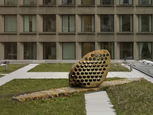 A large rock sculpture rests on a long strip of stone cylinders in a‬ ‭greenspace. The rock has a series of holes removed from its core, with the‬‭ cylinders that were removed making up the strip it rests on. There is a pathway behind the sculpture and building tops surrounding the open space.