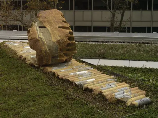 A large rock sculpture rests on a long strip of stone cylinders in a‬ ‭greenspace. The rock has a series of holes removed from its core, with the‬‭ cylinders that were removed making up the strip it rests on.