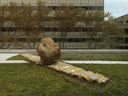 A large rock sculpture rests on a long strip of stone cylinders in a‬ ‭greenspace. The rock has a series of holes removed from its core, with the‬‭ cylinders that were removed making up the strip it rests on. There is a pathway behind the sculpture and building tops surrounding the open space.