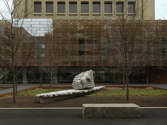 A large rock sculpture rests on a long strip of stone cylinders in a‬n open space. The rock has a series of holes removed from its core, with the‬ cylinders that were removed making up the strip it rests on. Trees, a pathway,‬ ‭and a gray building reside in the background.‬ ‭