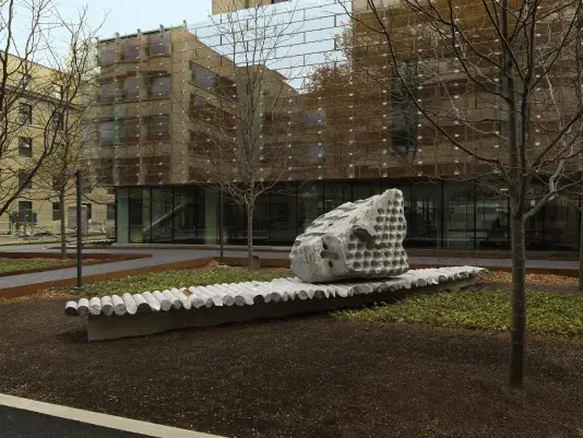 A large rock sculpture rests on a long strip of stone cylinders in a‬n open space. The rock has a series of holes removed from its core, with the‬ cylinders that were removed making up the strip it rests on. Trees, a pathway,‬ ‭and a gray building reside in the background.‬ ‭
